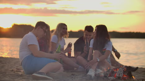 The-group-of-young-people-is-sitting-in-around-bonfire-on-the-sand-beach.-They-are-talking-to-each-other-and-drinking-beer-at-sunset.